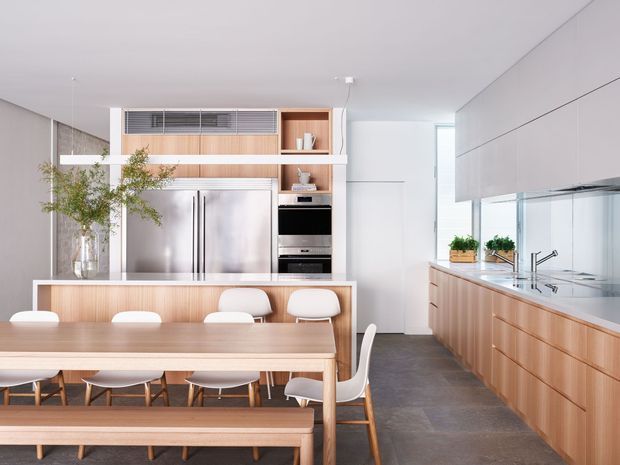 Bright contemporary open-plan kitchen and dining area with extensive light timber cabinetry, a large island with integrated seating, stainless-steel appliances, a long timber dining table and bench, and white dining chairs. The space features clean white walls and ceiling, pale timber joinery, dark stone-look flooring, and a mirrored or reflective splashback along the kitchen workbench.