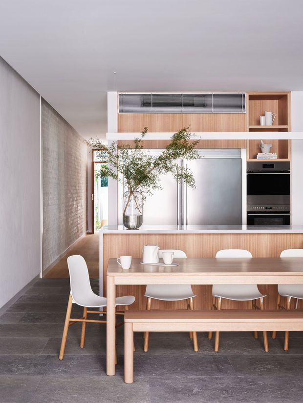A refined open-plan kitchen and dining area with a long timber dining table, matching bench, and white upholstered dining chairs. The kitchen features light timber cabinetry, stainless-steel appliances, open shelving, and a broad island or counter. A textured grey wall and dark stone-look flooring contrast with the warm timber joinery, while indoor greenery adds a natural accent. The space is bright, minimal, and carefully detailed.