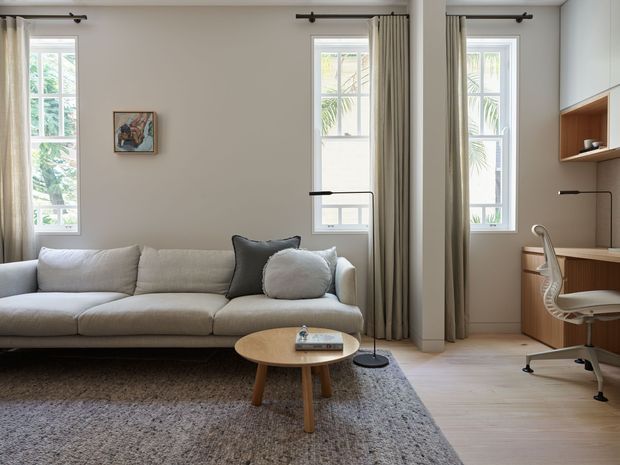 A bright, refined living room with a neutral upholstered sectional sofa, round timber coffee table, textured grey rug, and a slim floor lamp. Tall traditional-style windows with full-length curtains bring in abundant daylight. A compact built-in timber-toned home office with open shelving and a modern task chair occupies the right side of the room. Pale walls and light timber flooring create a calm, contemporary interior.
