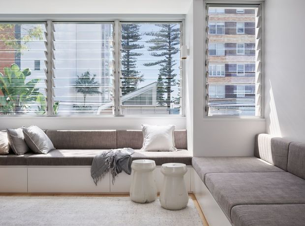 Bright, compact contemporary living room with a built-in window seat and an L-shaped grey upholstered sectional arranged around a low light rug. Large white-framed louvred windows provide abundant daylight and views of neighbouring apartment buildings and trees. Two small white side tables and a draped grey throw add simple furnishings, while integrated white cabinetry runs beneath the window seat.