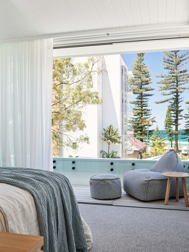 Bright coastal bedroom opening onto a balcony or terrace, with a partially visible bed in the foreground, sheer white curtains, grey textured bedding, a boucle lounge chair and ottoman, and a small round timber side table. The room has a white-lined ceiling, light carpet, and full-height opening with a glass balustrade. Through the opening are tropical landscaping, neighbouring buildings, and a glimpse of blue ocean under a clear sky.