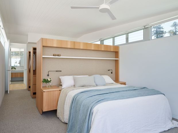 Bright, compact bedroom with a built-in timber bedhead and integrated bedside cabinetry. The room has a white panelled ceiling, high clerestory-style windows, neutral carpet, simple black wall-mounted reading lights, and layered white and pale blue bedding. A ceiling fan and an open passage toward a bathroom or dressing area are visible.