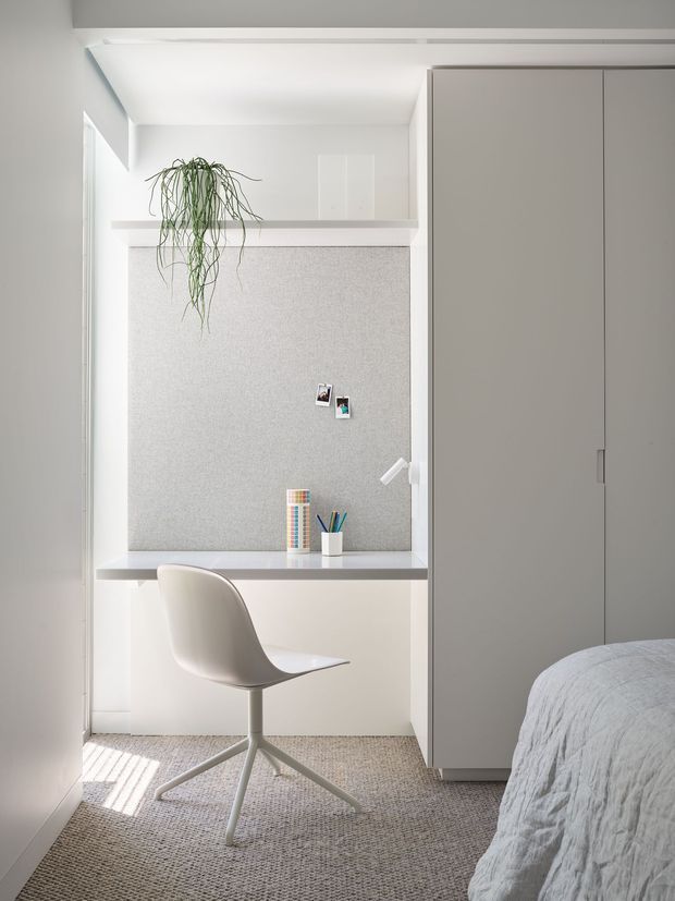 Bright, compact bedroom workspace with a built-in white desk and pinboard niche, a simple swivel chair, tall integrated wardrobe cabinetry, and part of a bed visible in the foreground. The room has white walls and joinery, a pale textured pinboard, soft grey carpeting, and minimal decor including a hanging plant and small desk accessories.