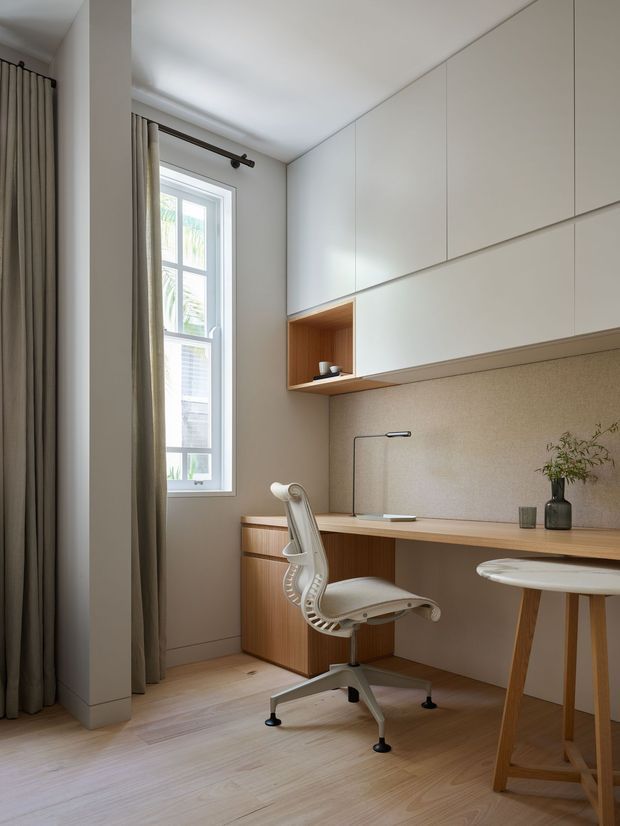 A compact, refined home office with a built-in timber desk, extensive white overhead cabinetry, an open timber display niche, and a light ergonomic task chair. A tall window with full-length neutral curtains brings in natural light, while a slim desk lamp, vase, and small side table complete the calm workspace. Pale timber flooring and warm wood joinery create a soft, understated interior.