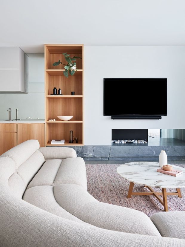 A bright, contemporary open-plan living room with a large curved cream upholstered sectional in the foreground, a round marble coffee table, and a wall-mounted television. The room features a grey stone fireplace surround and hearth, integrated timber shelving with decorative objects and a plant, and a compact timber-and-white kitchen visible to the left. The palette is light and neutral with warm timber accents and soft textured furnishings.