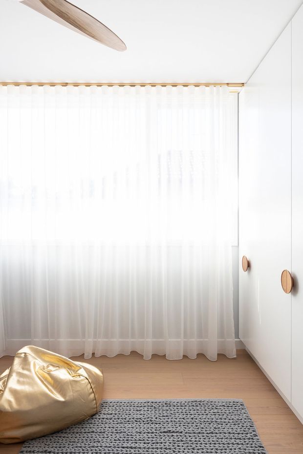 Bright, minimalist bedroom corner with full-height sheer white curtains, pale timber flooring, a large grey textured rug, a metallic gold beanbag, and white built-in wardrobes with rounded timber-toned handles. A sculptural timber ceiling fan is partially visible, and the room is filled with soft natural light.