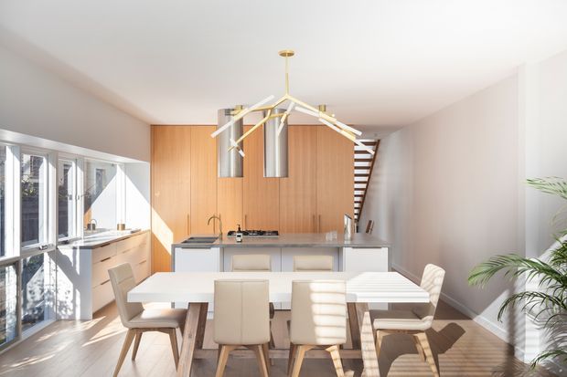 Bright contemporary open-plan kitchen and dining area with a central island, warm timber wall cabinetry, pale stone surfaces, a linear dining table, upholstered dining chairs, and a sculptural brass-and-white pendant light. Full-height glazing brings in strong sunlight, while a partially visible open-riser staircase leads upward at the rear.