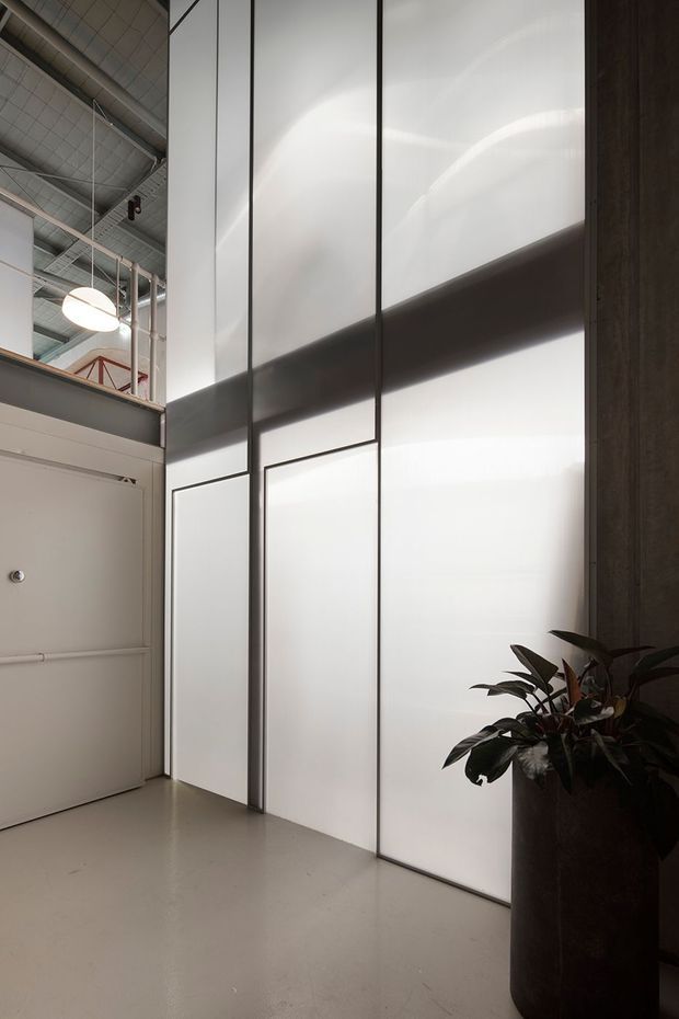 A tall, minimalist interior corridor or entry area in an industrial-style building. Large translucent white wall panels are divided by slim dark vertical and horizontal framing, creating a luminous feature wall. The space has a smooth light-grey floor, an exposed high ceiling with visible services and suspended lighting, a plain white door or partition at left, and a dark planter with broad-leaf greenery in the foreground.
