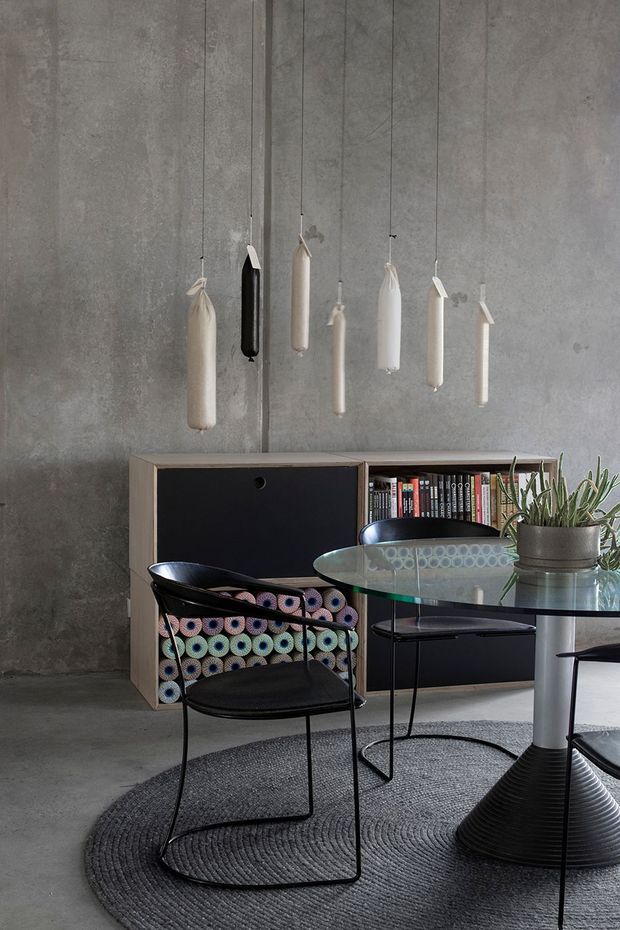 A moody, minimalist dining and living area with a raw grey concrete wall and floor. A low timber-and-black storage unit holds books and rolled textiles, while a round glass dining table with a sculptural black pedestal base is surrounded by slim black chairs. Several elongated hanging pendant lights create a sculptural focal point, and a potted plant adds restrained greenery. A dark textured round rug anchors the furniture.