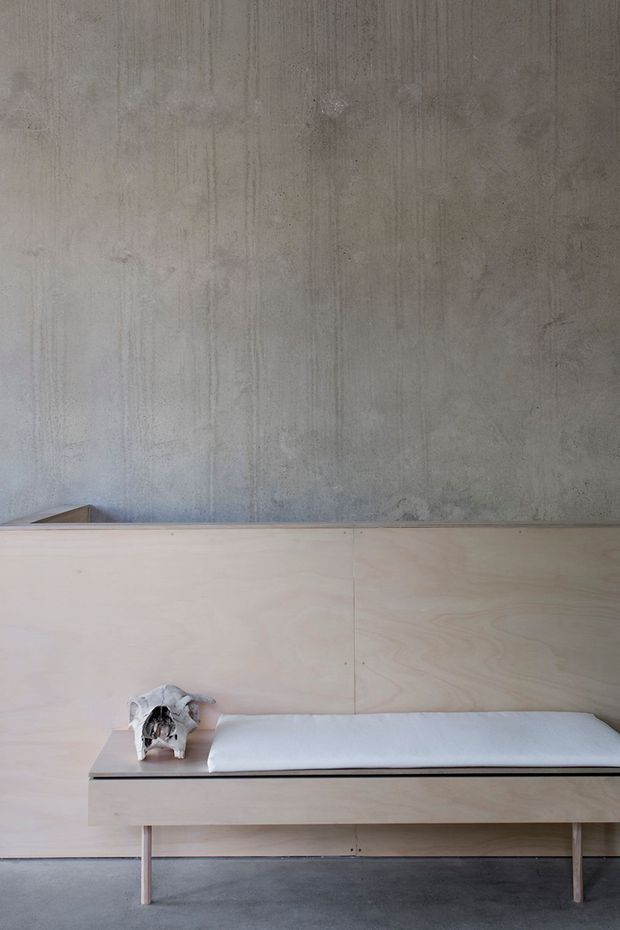 Minimalist interior detail featuring a pale plywood built-in bench or daybed against a raw grey concrete wall. The bench has a slim black-edged top, white cushion, and simple timber legs, with a small sculptural object placed at one end. A dark grey concrete floor and restrained palette create a quiet, utilitarian composition.