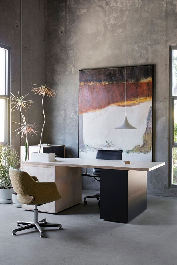 A moody contemporary home office with a concrete-finished interior, polished concrete floor, tall dark-framed windows, and a minimalist pale timber and black desk. Two office chairs sit beneath a sculptural pendant light, with a large abstract artwork and sparse potted plants adding visual interest.