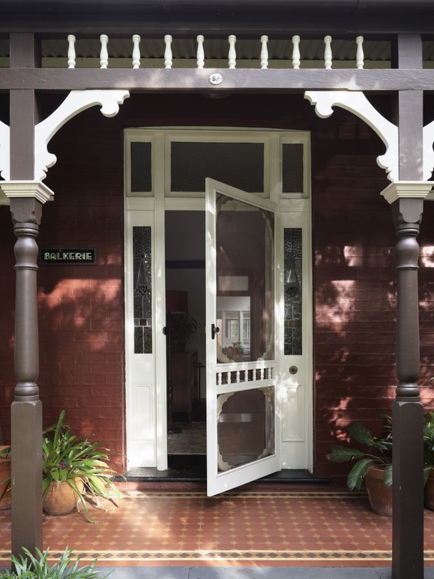 Front entrance of a heritage-style brick building with an ornate covered verandah, dark timber posts, decorative fretwork, turned balusters and a white glazed timber door standing open. Patterned terracotta and cream tiled paving, potted plants and a small entry plaque add character to the welcoming porch.