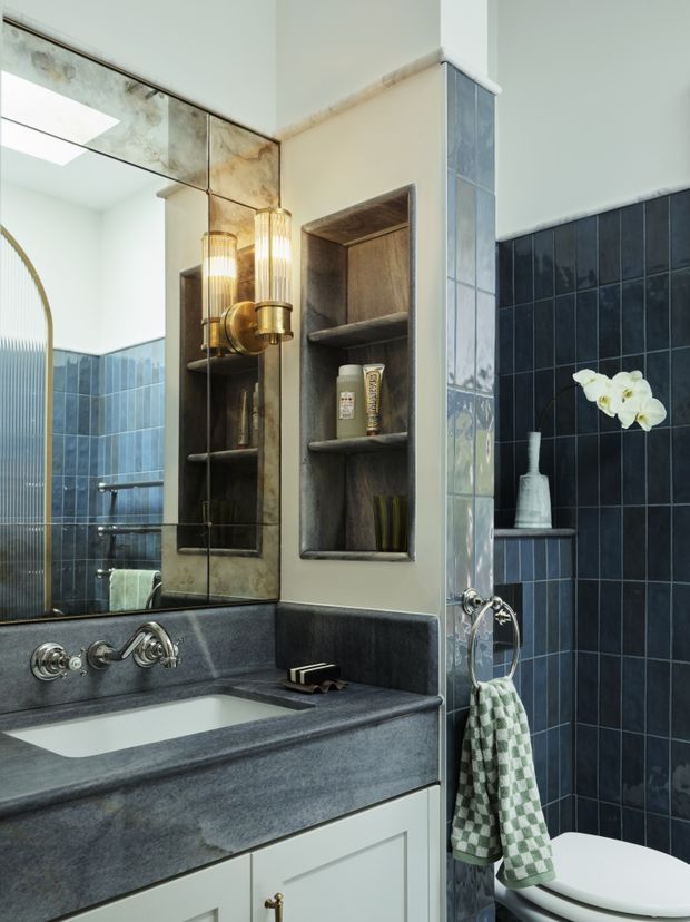 A compact, richly detailed bathroom featuring a dark blue tiled shower and wall, a deep grey stone vanity benchtop with integrated white basin, pale cabinetry, mirrored wall, recessed dark shelving, polished metal fixtures, and a warm vertical pendant light. A partial bathtub and toilet are visible, with a decorative orchid adding contrast.