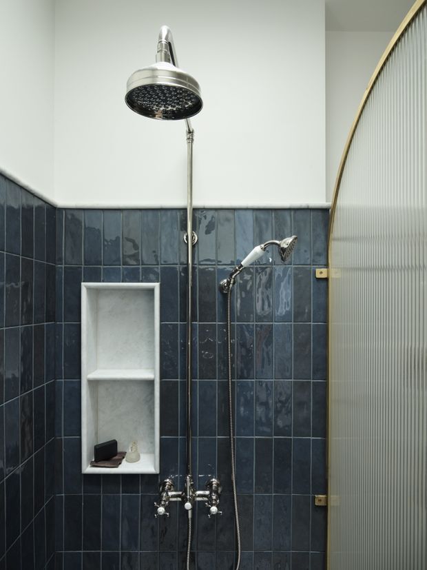 A compact shower area with dark blue glossy wall tiles covering the lower walls, white upper walls, and exposed polished chrome shower fixtures including a large rain showerhead and handheld shower. A recessed white tiled niche provides storage, while a curved ribbed translucent glass screen frames the right side.