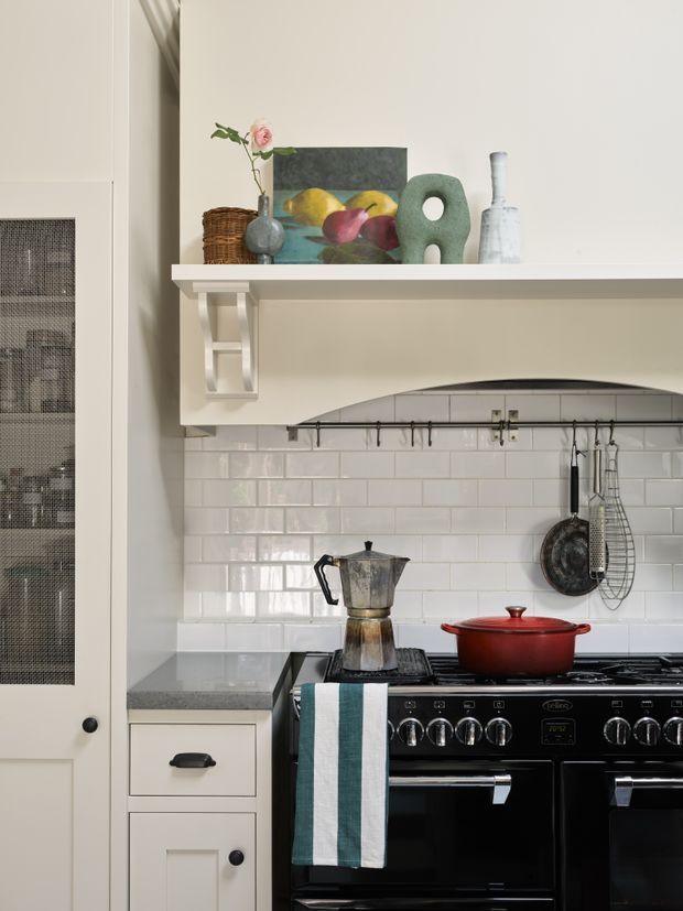 A detailed close-up of a traditional-style kitchen range area with a black freestanding cooker, dark stone countertop, white glossy subway-tile splashback, and a painted mantel-style range hood with decorative bracket. Open shelving above displays pottery, a basket, artwork, and a vase. Cooking utensils hang from a rail, with a moka pot and red Dutch oven on the stovetop.