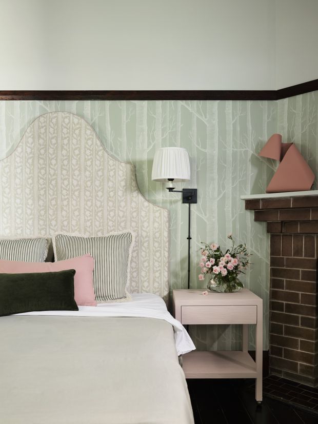 A styled bedroom corner with a bed featuring a scalloped upholstered headboard, layered striped and solid cushions, pale bedding, a blush-pink bedside table, wall-mounted lamp, floral arrangement, and dark brick fireplace. The walls combine pale green botanical wallpaper with a patterned inset behind the bed, accented by dark timber trim and a sculptural pink object on the mantel.