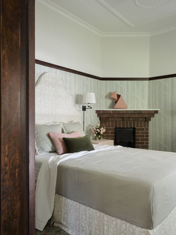 A cozy, traditionally detailed bedroom viewed through a dark timber doorway. The room features a made bed with a patterned upholstered headboard and bed skirt, pale bedding, blush and olive cushions, a brick fireplace with a timber mantel, patterned pale wallpaper, dark timber trim, decorative ceiling mouldings, a wall-mounted lamp, and a small sculptural object.
