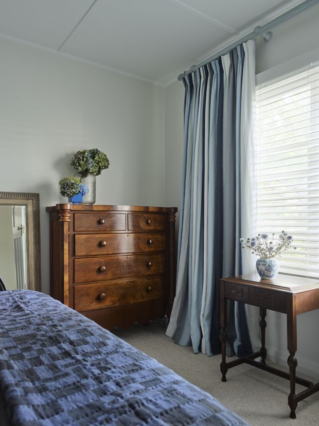 A refined traditional bedroom corner featuring a dark timber chest of drawers, a narrow side table, blue-and-white striped floor-length curtains, plantation shutters, patterned blue bedding, and floral arrangements. The room is softly lit with a calm, classic atmosphere.