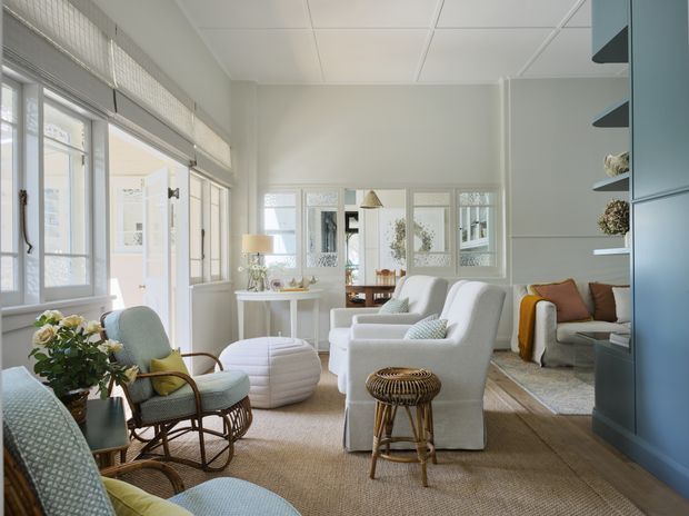 Bright, airy living room with large windows and glazed doors, white upholstered seating, wicker and rattan furniture, a textured natural-fibre rug, and pale blue built-in shelving and cabinetry. The room has a relaxed coastal character with abundant daylight and soft neutral finishes.