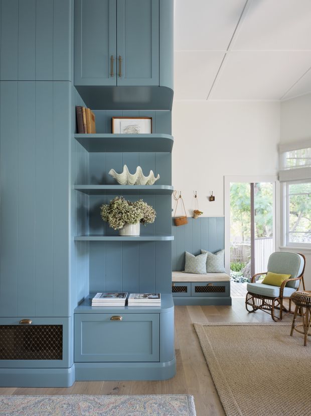 A light-filled, carefully detailed living room with a built-in blue-grey cabinetry and shelving wall, including upper cupboards, open display shelves, and lower storage. The joinery incorporates brass pulls and a dark woven grille panel. A window seat with cushions sits beneath a half-height blue panelled wall, while a rattan lounge chair and side table occupy the foreground. Pale timber flooring, layered woven rugs, white ceilings, and garden views through the glazed doors create a calm coastal character.