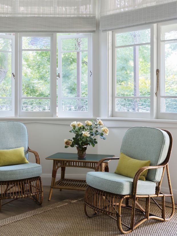 Bright, serene sitting area with two woven rattan lounge chairs upholstered in pale blue fabric and accented by mustard-yellow cushions. A small dark-top wicker table with a vase of flowers sits between the chairs. White-trimmed multi-pane windows with woven Roman blinds overlook leafy greenery, while a natural textured rug covers the floor.
