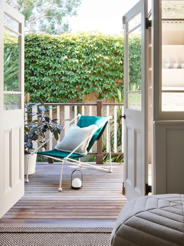 Bright view from a bedroom through open glazed French doors onto a compact timber deck or balcony. The outdoor space has a turquoise sling chair with a cushion, a potted plant, timber decking, and a white-and-brown railing backed by a dense leafy privacy screen. The bedroom foreground includes a grey upholstered bed and neutral patterned rug. Built-in shelving with decorative panels is visible at right.