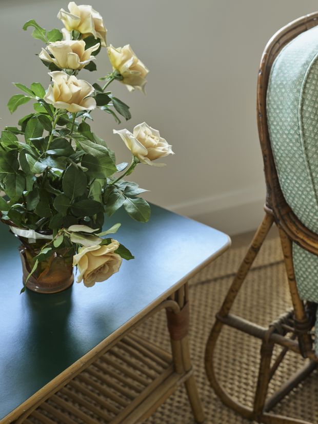 Close-up interior vignette featuring a dark green rectangular table with rounded edges, a glass vase of cream roses, and a woven rattan chair with a pale green upholstered back. The scene has soft natural light, muted neutral walls, and a warm, refined vintage-inspired feel.