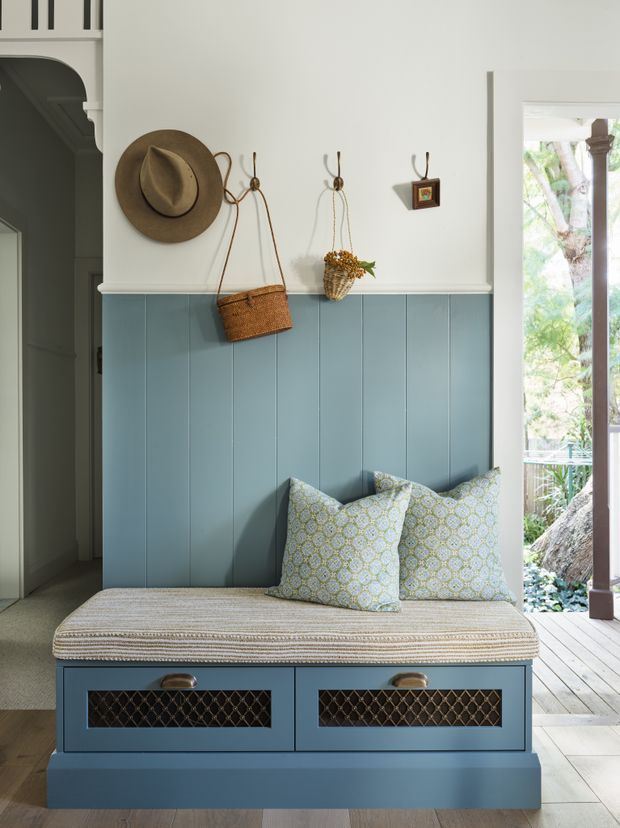 A bright, refined entryway featuring a built-in blue painted timber bench with upholstered seat cushion and patterned pillows. The wall has blue vertical panelling below a white upper section, with hooks holding a hat, woven bag, and small basket. A decorative arched opening is visible to the left, while glazed doors on the right open to a leafy outdoor deck.