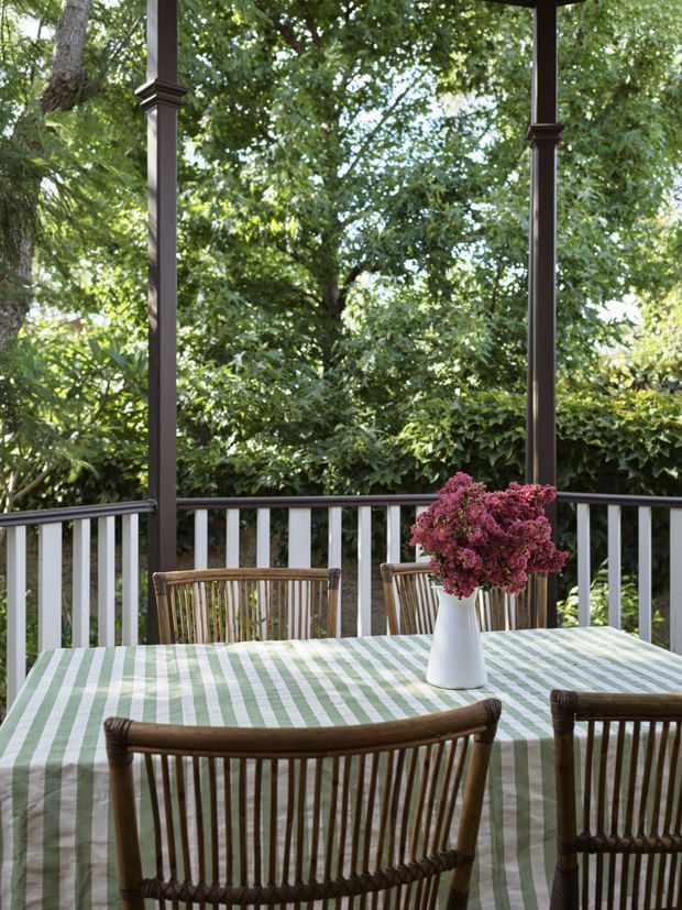 A covered outdoor dining veranda with dark timber posts, white balustrading, and a dining table covered in a green-and-white striped cloth. Woven timber dining chairs surround the table, which holds a white vase of pink flowers. Dense leafy garden vegetation fills the view beyond the railing, creating a shaded, garden-facing setting.