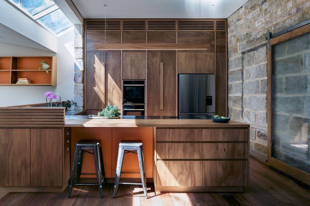 A warm, contemporary kitchen with extensive dark timber cabinetry, a central island with seating, integrated appliances, and a stone masonry wall. Natural light enters through a large skylight, while open shelving and a deep terracotta-toned backsplash add colour and character.