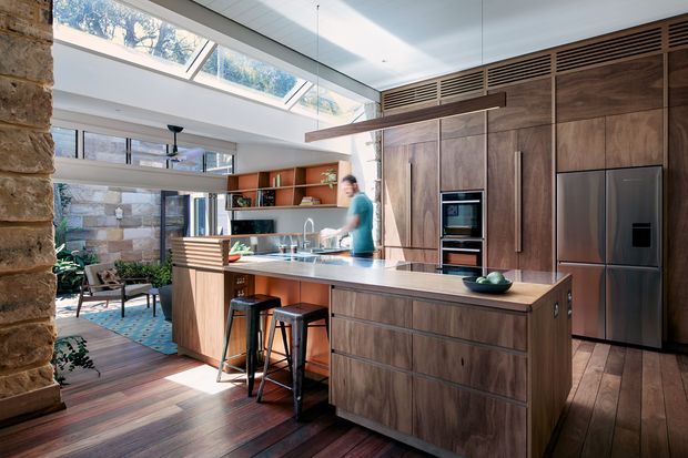 A bright, contemporary open-plan kitchen with a large timber island, integrated dark wood cabinetry, stainless-steel appliances, and a linear pendant light. Skylights and clerestory glazing provide abundant natural light, while a stone feature wall and adjoining lounge area create a warm indoor-outdoor feel.