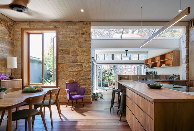 Bright open-plan kitchen and dining area in a renovated stone-walled home. The space features a long timber kitchen island with integrated cooktop and seating, extensive timber cabinetry, exposed sandstone walls, timber flooring, a white-lined ceiling, and large clerestory and skylight-style glazing. A dining table, mid-century-inspired chairs, and a purple armchair occupy the adjacent dining zone, with abundant natural light and greenery visible through the windows.