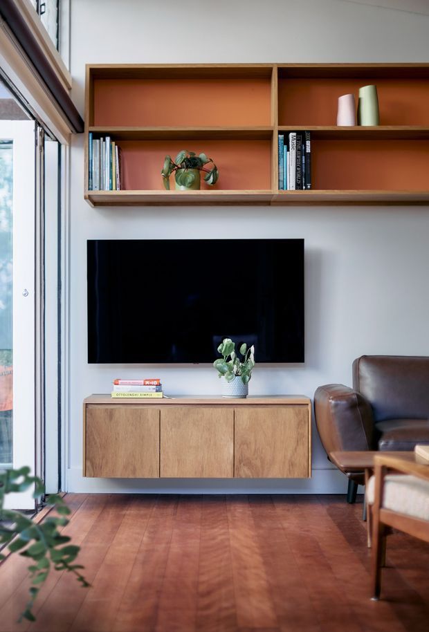 A compact, warm living room vignette featuring a wall-mounted television, a floating timber media console, and an open timber display shelf with a terracotta-painted backing. A leather armchair sits to the right, while large glazing on the left brings in daylight. The room has timber flooring, white walls, and a mid-century-inspired palette of natural wood, terracotta, and brown leather.