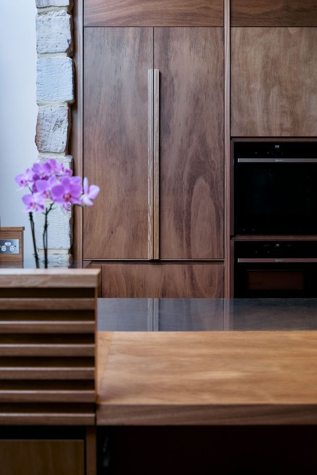Close-up view of a warm, richly timber-lined kitchen featuring tall integrated cabinetry, long vertical handles, built-in black appliances, and a timber island with a dark stone benchtop. A rough pale masonry wall and purple orchid add textural and colour contrast.