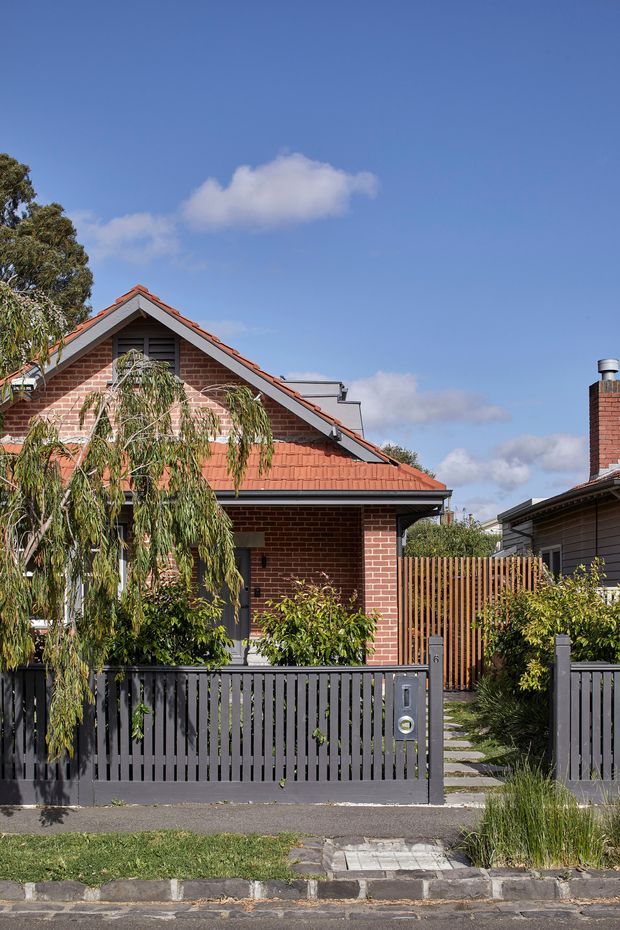 Street-facing view of a compact brick suburban house with a red terracotta tiled gable roof, dark grey fascia and front fence, timber pedestrian gate, landscaped planting, and a paved path leading to the entrance. A neighbouring weatherboard building and chimney are visible at the right edge under a clear blue sky.