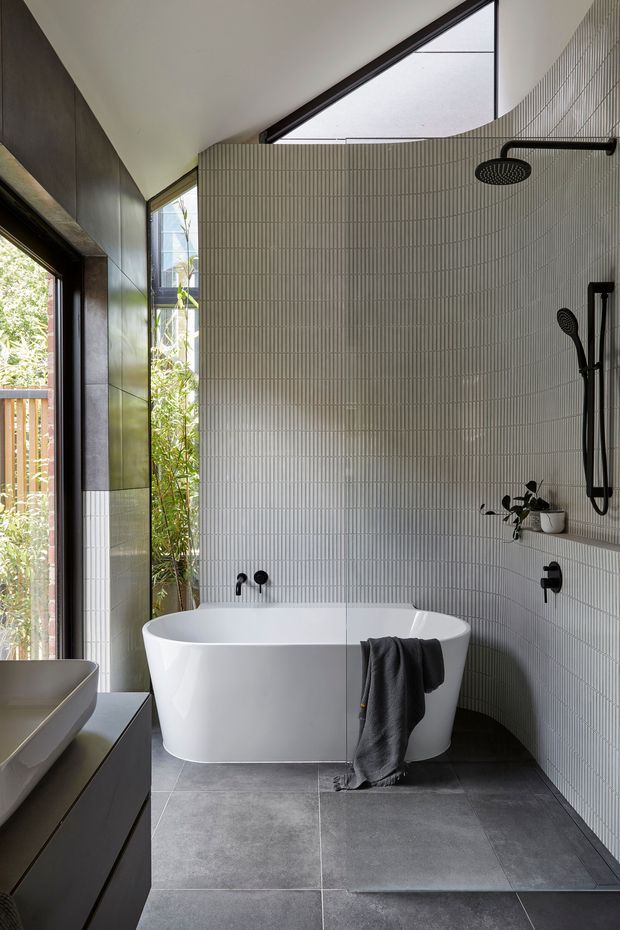 A contemporary bathroom with a freestanding white bathtub, large-format dark grey floor tiles, and vertically ribbed light grey wall tiles. Matte black shower fixtures, taps, and hardware contrast with the pale surfaces. Full-height glazing brings in natural light and views of greenery, while dark built-in cabinetry and a partial vanity are visible at left. The room has a high, angular ceiling with a clerestory-like glazed section.