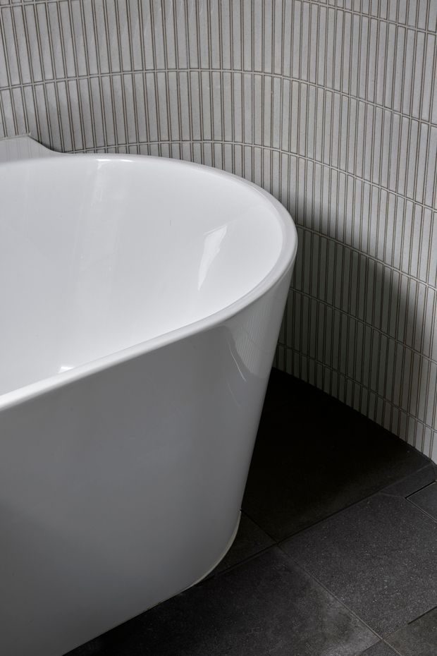 Close-up view of a contemporary bathroom featuring a freestanding white oval bathtub against a wall of narrow vertically arranged light grey tiles. Large dark grey floor tiles create a strong contrast with the glossy tub and pale wall finish.