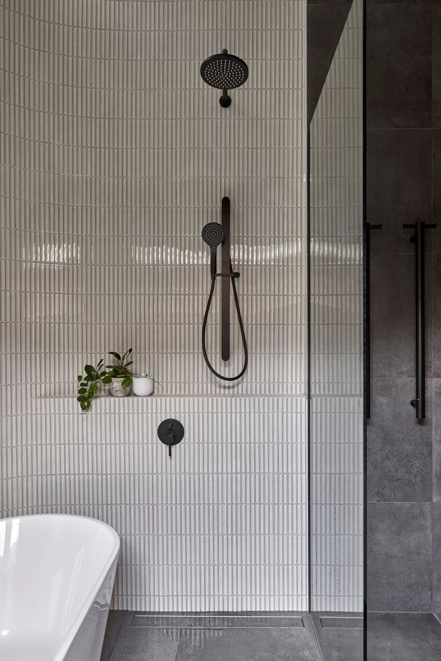 A contemporary bathroom with a walk-in shower, featuring vertically ribbed light grey wall tiles, dark charcoal floor and adjacent shower-wall tiles, matte black shower fittings and glass shower screen. The corner of a white freestanding bathtub and a small recessed shelf with potted plants are visible.