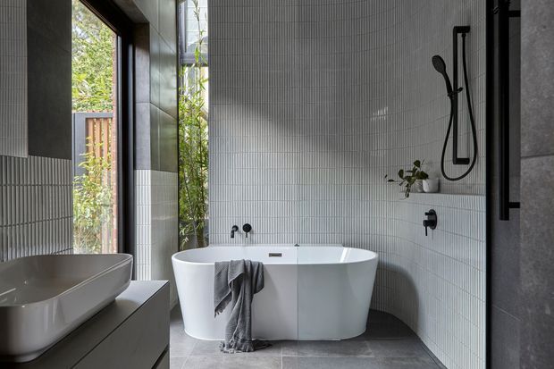 A contemporary bathroom with a freestanding white bathtub positioned against a curved wall clad in narrow white vertical tiles. A separate white vessel basin sits on a dark built-in vanity at left. The room includes a black-framed shower enclosure with a wall-mounted black hand shower, dark grey floor tiles, large black-framed windows, and natural greenery visible outside.