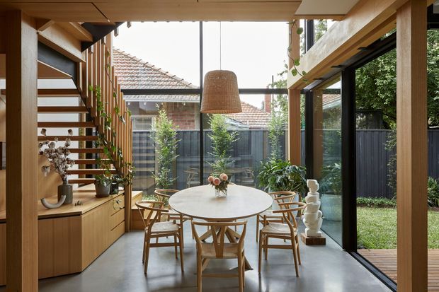 A light-filled contemporary dining area with a round table and timber chairs, framed by extensive floor-to-ceiling glazing opening to a small courtyard. Warm timber ceiling, shelving, cabinetry and an open-riser staircase surround the space, with a woven pendant light and indoor plants adding natural texture. Neighbouring tiled roofs and a dark privacy fence are visible outside.