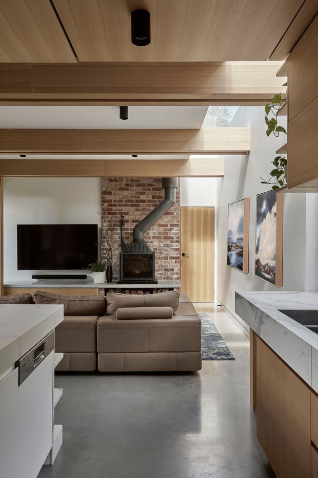 Open-plan living and kitchen interior with a contemporary timber-lined ceiling and integrated cabinetry. A large sectional sofa faces a television and a compact wood-burning stove set against an exposed red-brick feature wall. The kitchen includes a white marble-look benchtop and warm timber cabinetry, while a skylit passage and timber door bring natural light into the space. The room combines polished concrete flooring, timber joinery, exposed brick and restrained modern furnishings.