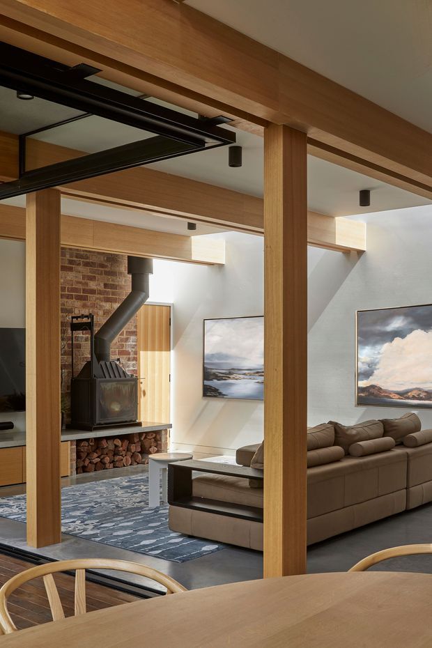 A warm, open-plan interior viewed from a dining area, with a timber dining table and chairs in the foreground. Timber posts and beams frame a living room containing a large brown sectional sofa, patterned rug, and a brick fireplace with a black wood-burning stove and stacked firewood. White walls, timber detailing, recessed spotlights, artwork, and dark flooring create a refined contemporary-rustic atmosphere.