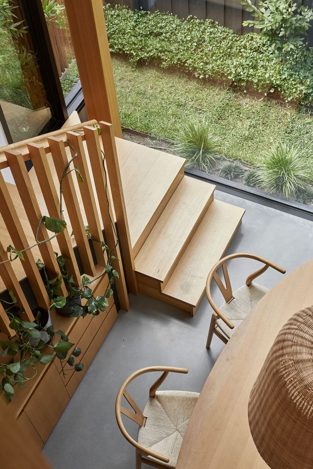 A warm, compact interior with a timber dining table and woven-seat wooden chairs beside a set of built-in timber steps. Vertical timber slats form a balustrade and planter feature, while full-height glazing looks onto a small landscaped courtyard. The space has a refined natural aesthetic with abundant greenery and a smooth grey floor.