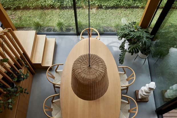 Overhead view of a warm, light-filled dining area with a large oval light-wood dining table, woven pendant light, and curved timber dining chairs. Floor-to-ceiling glazing opens onto a planted garden, while timber stairs and a slatted timber balustrade are visible to the side. The room has a contemporary, nature-connected feel with concrete flooring and abundant indoor planting.