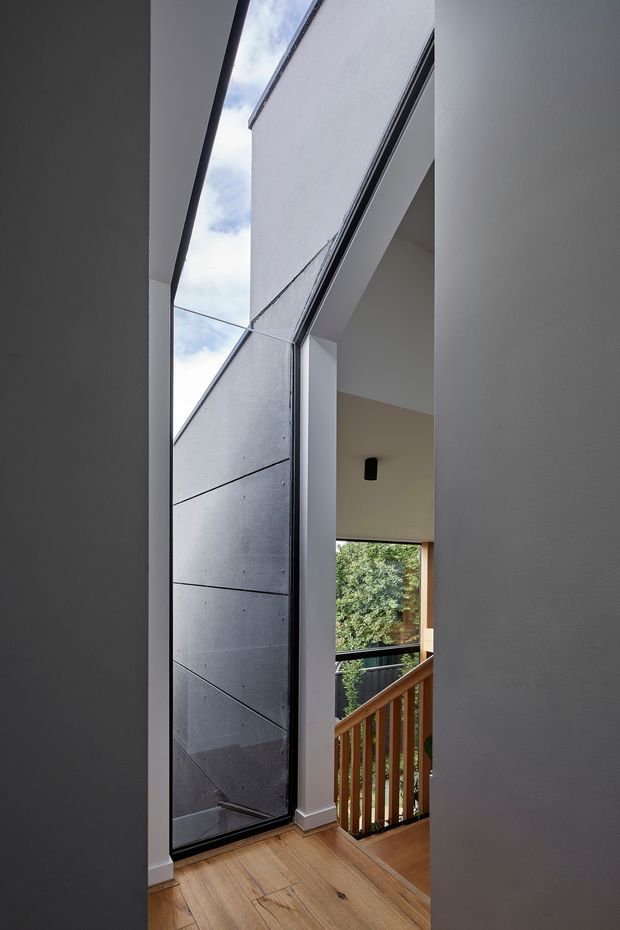 A narrow, vertically framed interior view of a modern stair landing or hallway. The space features a tall glazed opening or door with dark metal framing, pale plaster walls, warm timber flooring, and a timber stair balustrade. A large angular void brings in daylight and views of greenery outside, creating a strong connection between the circulation space and the exterior.