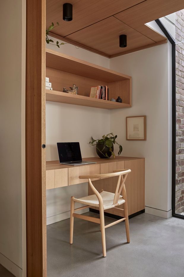 A compact built-in home office nook viewed through a timber-framed opening. It features a warm timber-lined ceiling with recessed black cylindrical downlights, open timber shelving, a timber credenza with integrated drawers and doors, a laptop workspace, a light timber chair, and a potted plant. A large glazed opening beside exposed brick brings in natural light, while the polished concrete floor and soft off-white walls create a refined, warm contemporary interior.