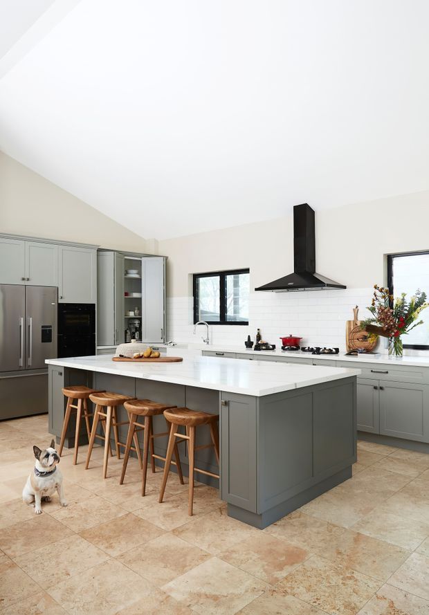 Bright, spacious kitchen with a large grey island, white benchtops, integrated seating for four, grey shaker-style cabinetry, stainless-steel appliances, a black rangehood, white tiled splashback, and warm beige stone-look floor tiles. Large windows provide natural light, with decorative timber boards and flowers on the rear bench. A small dog is visible on the floor.