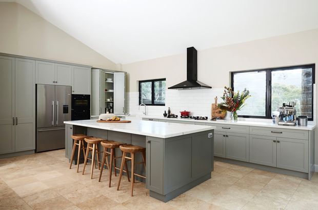 Bright, spacious contemporary kitchen with a large central island, integrated seating, grey raised-panel cabinetry, stainless-steel appliances, pale stone benchtops, tiled flooring, black rangehood, and generous windows.