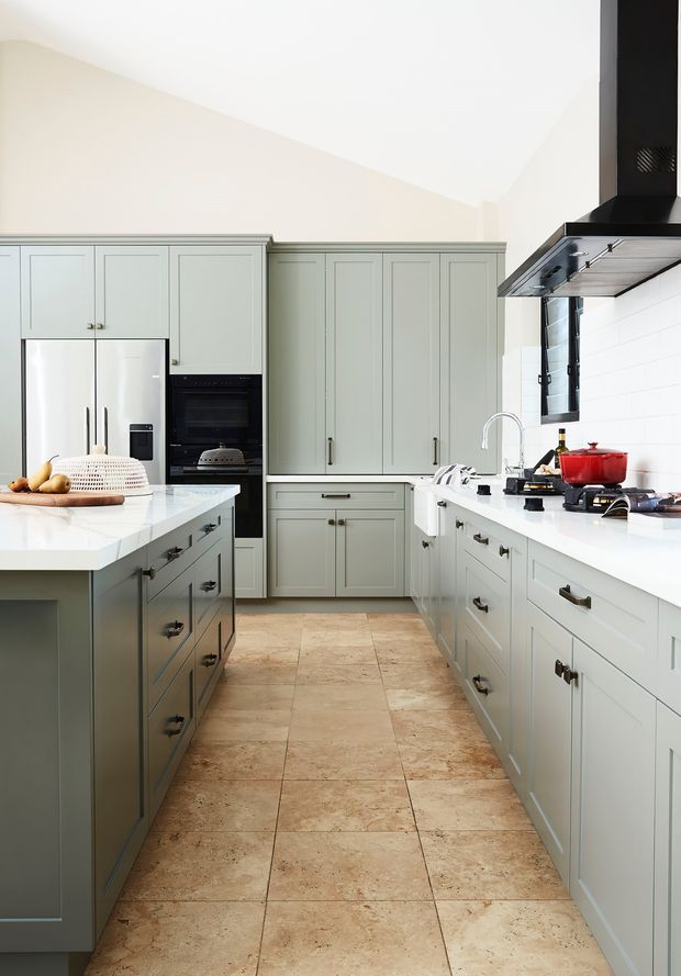 Bright, spacious classic kitchen with pale sage-green shaker cabinetry, white stone countertops, a central island, integrated stainless-steel refrigerator and black oven, a large black rangehood, and a white subway-tile splashback. Large beige stone-look floor tiles and black hardware provide contrast. The kitchen has a clean, refined, high-end residential feel.