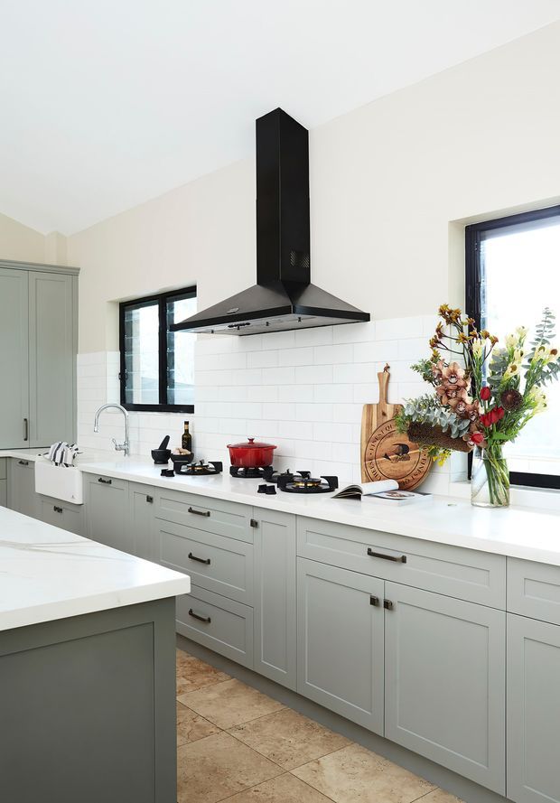 Bright, traditional-style kitchen with pale grey shaker cabinetry, white benchtops, white subway-tile splashback, black hardware and a prominent black rangehood. The room includes a large island, gas cooktop, farmhouse-style sink, dark-framed windows and warm stone-look floor tiles, with floral arrangements and timber accessories adding softness.